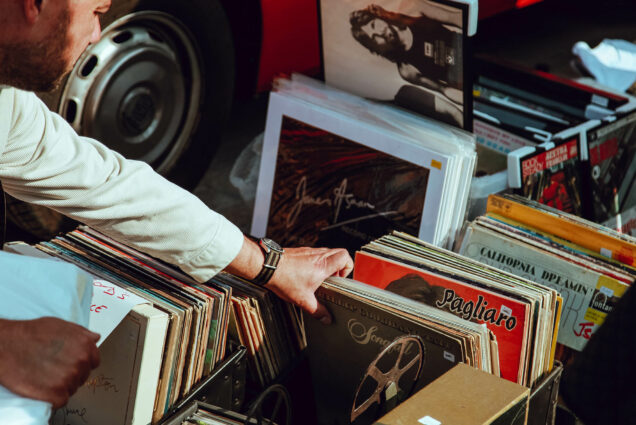 Photo: A white man's hand reaches for a folder of records. He is grabbing onto a handful of them. He is wearing a black wrist-watch and a white button-up shirt. There is a car in the background, with only the tires and the red bottom of the care showing.