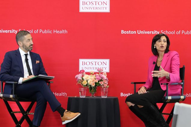 Photo: A red background with Boston University and Public Health text. Two people sit down on a stage, one a white man in a black suit and a white woman in a black outfit with a neon pink blazer. They sit between a small table with a black tablecloth over it.