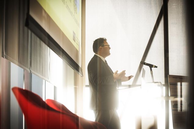 Photo: A man in a suit stands in front of a podium with a washed out white background and sun shining in through nearby windows