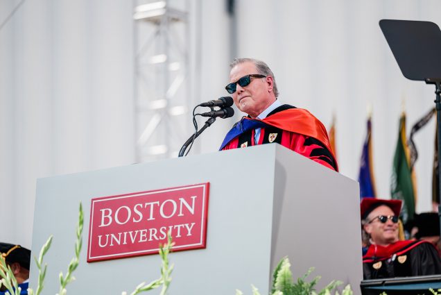 Photo: David Zaslav, a white man wearing a red ceremonial robe, black stole, and black sunglasses, speaks at a podium on stage with pointer fingers pointed up. Others wearing similar wear sit on either side of him.