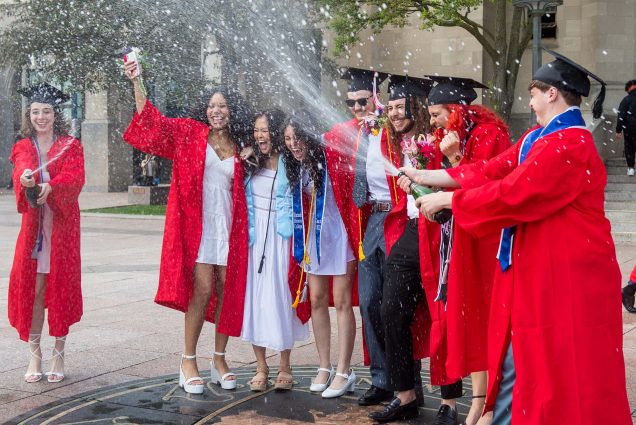 Photo: A group of students wearing BU scarlet red graduation gowns, black graduation caps, and formal wear laugh and smile as they stand on the BU Seal and spray newly opened bottles of champagnes into the air.