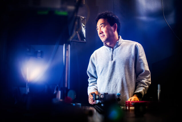 Photo: Engineering Professor, Dr. Ji-Xin Cheng, an Asian man wearing a grey pullover sweater, poses for a photo in his lab. A blue light gives the photo a cool-themed feel as Cheng looks to the left and stands in front of a table full of various mechanical engineering equipment.