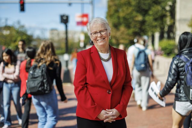 Photo: Portrait of Boston University Provost Jean Morrison on Commonwealth Ave. Asmiling white woman with short, silver white hair and wearing glasses, a white blouse, and red blazer poses with fingers clasped in front of her on a bustling sidewalk. Background and passerby are blurry.