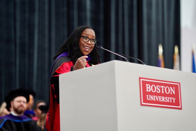 Photo: Ketanji Brown Jackson, a Black woman with long dreadlocks wearing a red ceremonial and purple stole smiles as she speaks into a white Boston University podium.