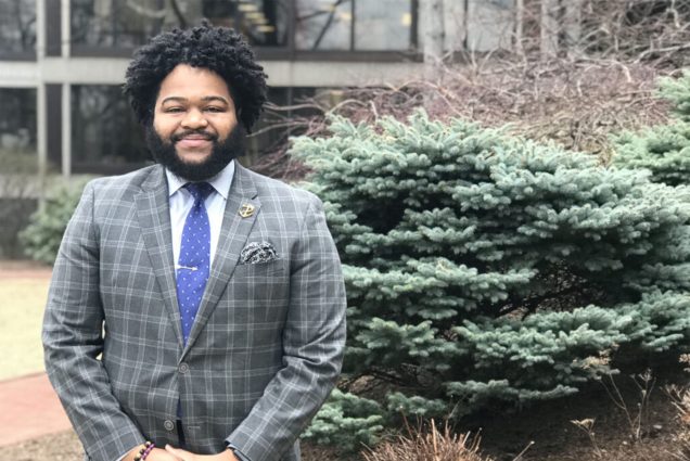 Photo: A black man with natural hair and a large beard wearing a patterend suit and blue tie stands in front of a coniferous tree with a smile on his face