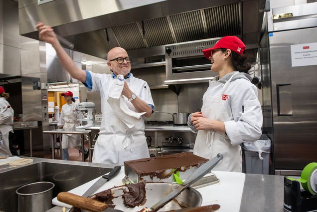 Photo: Chef Bill Yosses, an older white man wearing glasses and a white chef outfit, cleans off his ganache-spreading tool - a plastic ruler - with a flourish at the amusement of student Mara Clement, a younger white woman with dark curly hair tied back and wearing a red cap and white chef's outfit.