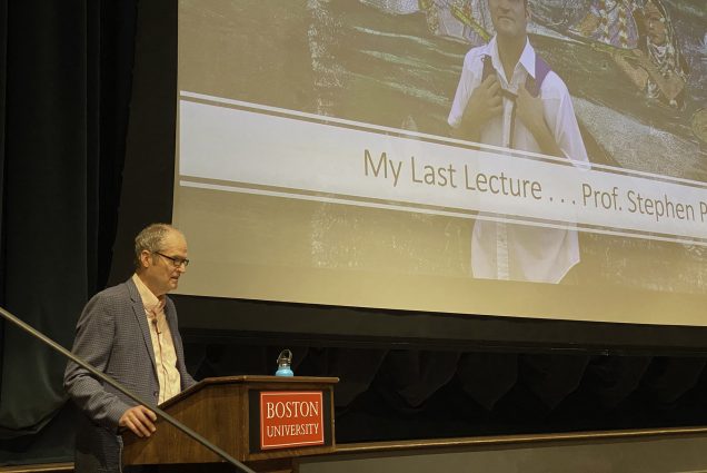 An older white man with dark gray hair and wearing glasses, a cream-colored collared shirt and grey blazer, reads from a podium as a large screen behind him displays a powerpoint that reads "My Last Lecture".