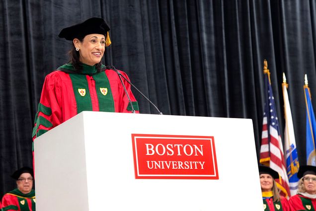 Photo: Rochelle Walensky, a tan woman wearing a red ceremonial graduation gown, black cap, and dark green stole, smiles as she gives a speech at a large white podium labelled with a red "Boston University" sign.