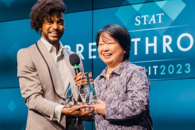 Photo: BU’s Xin Zhang (right) collects the STAT Madness 2023 All-Star award from Nicholas St. Fleur, a STAT general assignment reporter and associate editorial director of events. An Asian woman (right) with short black hair and wearing a purple collared shirt holds up a clear glass trophy with a Black man with brown hair in a curly poof and wearing a tan suit.