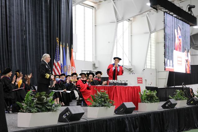 Photo: Sandro Galea (right) dean of the School of Public Health and Robert A. Knox Professor, applauds Admiral Rachel Levine, assistant secretary for health at the US Department of Health and Human Services. They stand on a enclosed stage--the dean in their ceremonial clothes and the admiral in their ceremonial outfit. Professors are sat in a line one the stage with multiple flags behind them. In the far right corner, there is a giant jumbotron.