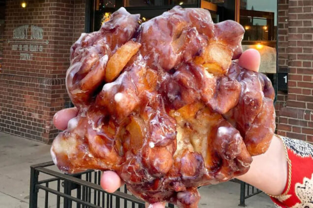 Photo: A hand holding a donut, freshly made, that is bigger than an average plam. Its an old-fashioned donut with a clear glaze. The setting is outside a brick-housed business.