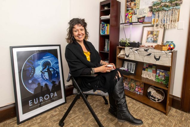 Photo of Romy Ruukel, Boston University director of the Shipley Center for Digital Learning & Innovation, in her office. A white woman with curly hair in a black blazer stis in an office filled with interesting trinkets and framed items.