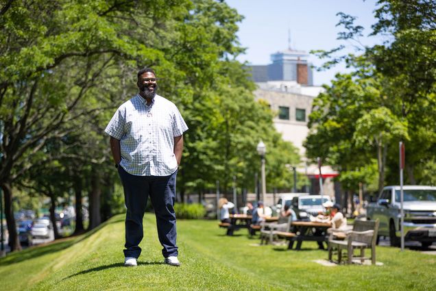 Photo: Anthony (Tony) Jack, a Black man wearing glasses, a light plaid collared shirt, and jeans, stands on a small grassy hill on a sunny day. Picnic tables filled with people can been in the blurry background behind him.