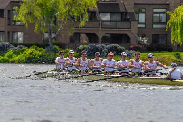 Photo: A group of men row a large boat with oars, they are all wearing matching red uniforms and tank tops