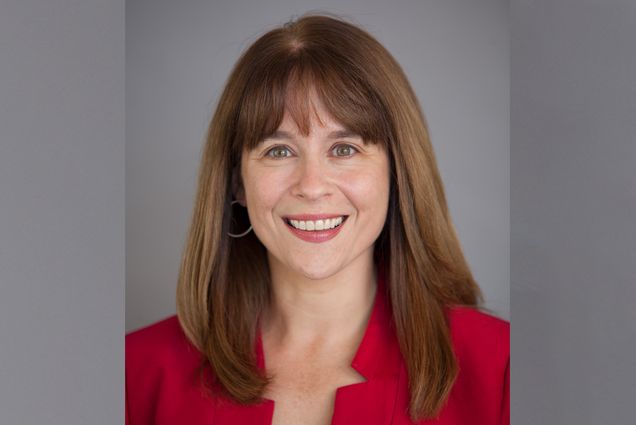 Photo: Headshot of Barbara Jones, a white woman with long, straight brown hair and wearing a red blouse. She smiles and poses in front of a grey backdrop.