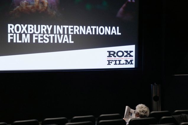Photo: A older person with white hair sits alone in the front of a movie theater. The text on screen reads "Roxbury International Film Festival"