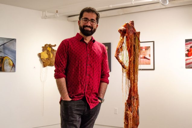 Photo: A person with glasses in a red shirt with a beard smiles at the camera in front of multiple pieces of art on canvas behind them