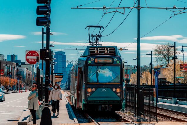 Photo: The MBTA Green B Line Trolley is shown on commonwealth Avenue. People stand on the sidewalk to the left as the T passes to the right. The Boston University and Boston city skyline can be seen i the distance on the sunny day.