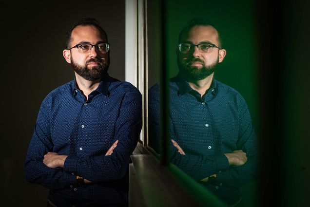 Photo: Gianluca Stringhini, a white man with short black hair and bear and wearing glasses and a long sleeved blue collared shirt, stands with arms crossed. He looks into a dark mirror as it shows his reflection.