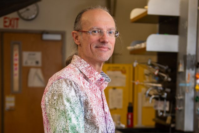 Photo of Mark Grinstaff, a middle-aged white man with glasses who wears a button-down shirt with a gradient, squiggly line pattern that fades from green and blue to red and pink. He smiles as he stands and leans against a cluttered lab table.