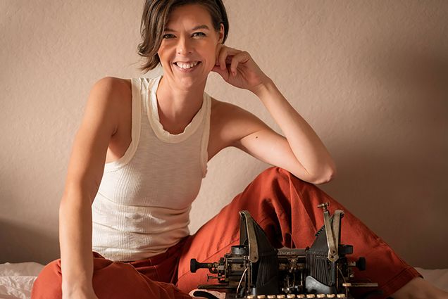 Photo: Jillian Abby, a white woman with short, brown hair and wearing a white tank top and orange-red casual pants, smiles and poses with one elbow resting on her knee. She sits on a white bed in front of a black, old school typewriter.