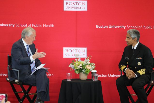 Photo: US Senator Edward Markey, an older white man wearing a black suit (left), and US Surgeon General Vivek Murthy, a South Asian man wearing a black naval uniform, sit in front of a red BU backdrop and are shown mid-discussion. A small black table with water bottles sits between them.