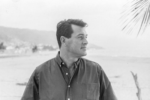 Black and white Photo: Rock Hudson, a white man wearing a long-sleeved collared shirt, looks to the right. Waves of large body of water from the beach shore can be seen in the blurred background behind him.