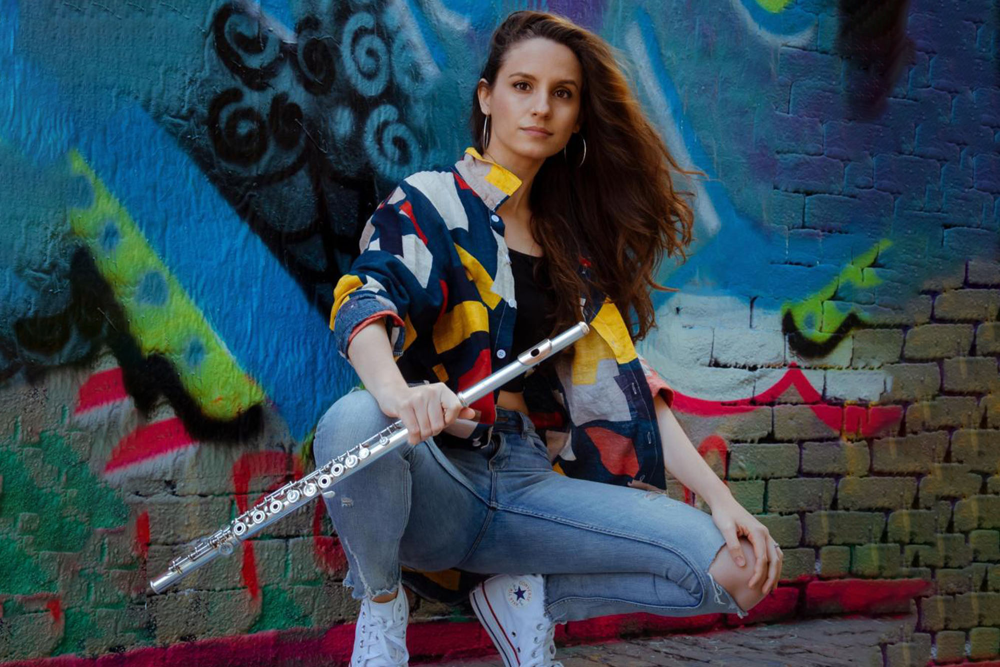 Photo: Sheila del Bosque, a white-presenting woman wearing a yellow, blue, and white collared shirt and jeans, squats and poses with silver flute in left hand. She poses in front of large blue mural.