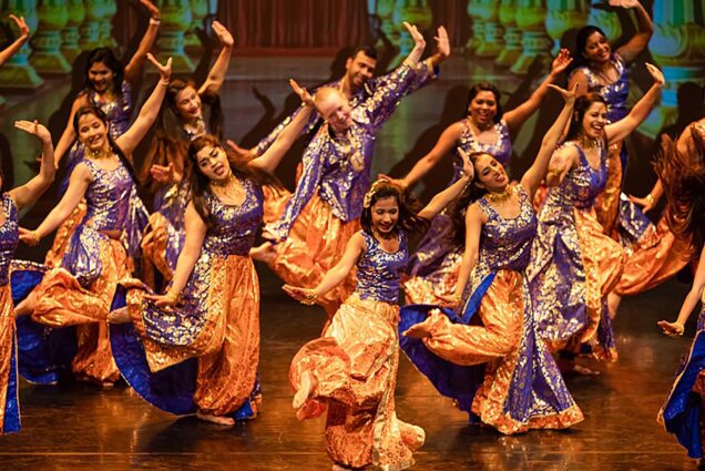 Photo: Colorful Bollywood dancers hit their dance move has the perform on stage. Their costumes consist of sparkly purple tank tops, with sparkly orange bottoms and a navy-blue lining on the pants. Most dancers have their hair down.