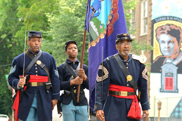 Photo: Three Black men dressed inn old military uniforms march in a Juneteenth parade. The young man in the middle holds a large, purple flag.