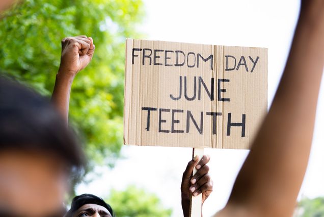 Photo: A cardboard sign that reads FREEDOM DAY JUNE TEENTH. On the left of the sign, a fist held up in solidarity by a Black individual. The individual holding the sign is also Black. The day is overcast and there is a blurred tree in the background.