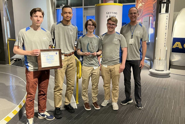 Photo: Five men in khakis pose for a photo after winning a competition. They all are wearing a gray top sponsored with the NASA logo on the left side. From left, a white man with short brown hair holds their certificate indicating their win. Next to him, a taller Black man with short brown hair stands next to a shorter white man in the middle, with floppy brown hair in glasses. The next two individuals are white men, one with glasses and blonde hair and the other is taller with balding hair.