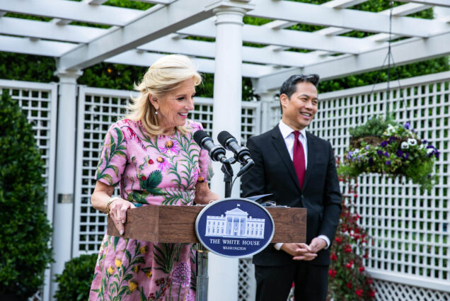 Photo: First Lady Jill Biden (left), an older white woman with blonde hair and wearing a pink and green floral dress, is shown speaking at a small wooden podium with the White House logo on the front. Director Richard Lui (right), a southeast Asian man with graying black hair and wearing a black suit, white collared shirt, and red tie, stands with hands clasped in front of him as he smiles to the unseen crowd.