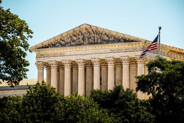 Photo: A sharp photo of the Supreme Court building. The foreground consists of the trees lining the front and on the right of the building, the American flag flys high. The day is bright and the sky is a blue clear.