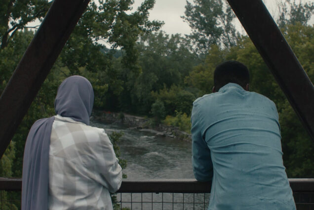 Photo: Two individuals stare with their backs facing the camera over a bridge. On the left, a woman with a hijab wearing a light-colored checkered shirt. On the right, a man in a blue button up.