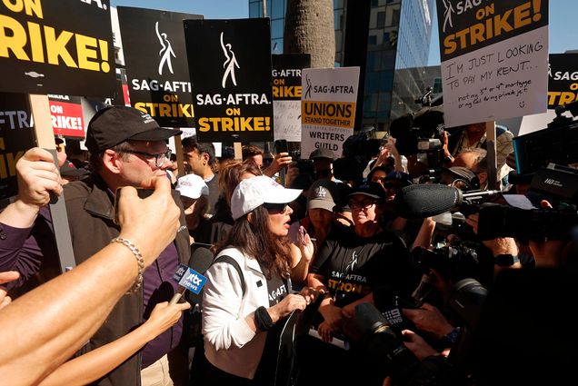 Photo: Fran Drescher, President of SAG, is swarmed by news outlets as she heads to a press call. She is wearing all white and stands in the middle while cameras and microphones surround her.