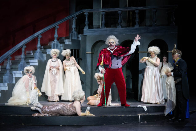 Photo: A man dressed in an intricate royal clown costume holds a sword drawn towards a person laying on the ground, as onlookers in white gowns gasp. The scene is part of an opera called Rigoletto