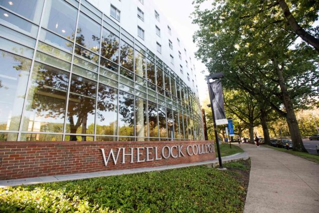 Photo: A angled shot of Wheelock College with a glass building and a brick sign with the metal words WHEELOCK COLLEGE on the outside. The day is bright and sunny.