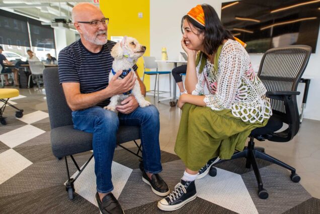Photo: Zoe Marsiglia, a young woman wearing a green dress and knitted white sweater, smiles as she sits next to, Lou Brum, an older white man wearing a navy blue and white striped shirt and jeans. A white cockapoo dog sits in his lap and looks excitedly at Marsiglia.