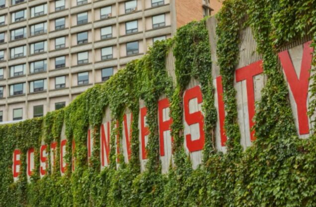 This is a photo taken on the Boston University campus. This landmark is seen on Nickerson Field and it is a cement wall that has large red lettering that spells out 'Boston University.' There is ivy grown up the cement wall, outlining the university's name. Behind the wall, there is a dorm building that can be seen. It is a tan, brick-looking building with many windows.