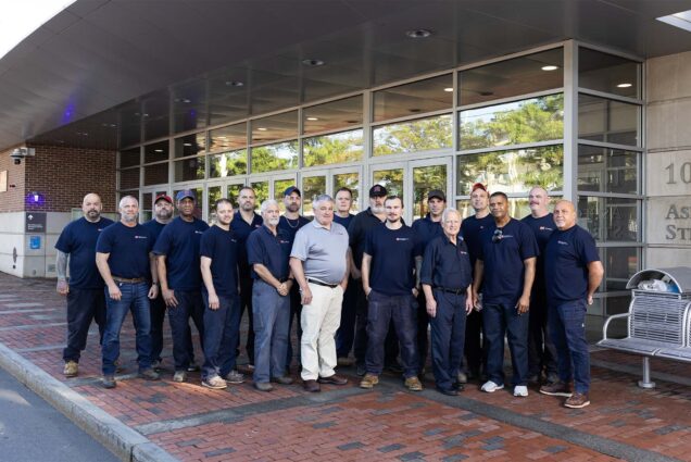 Photo: A team of HVAC technicians at Boston University pose for a photo in front of their headquarters. They are all wearing dark blue uniforms, except for the boss who is wearing a collared shirt in the center of the photo.