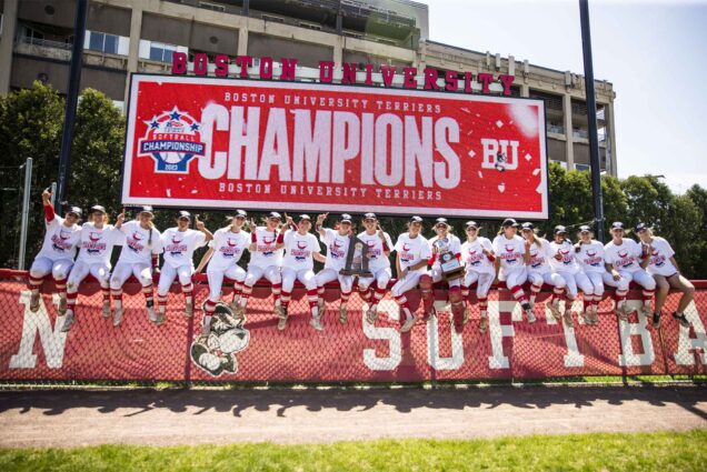 Photo: The BU Women's softball team sits atop the outfield fence with a banner reading "Champions" after winning the Patriot League in 2023