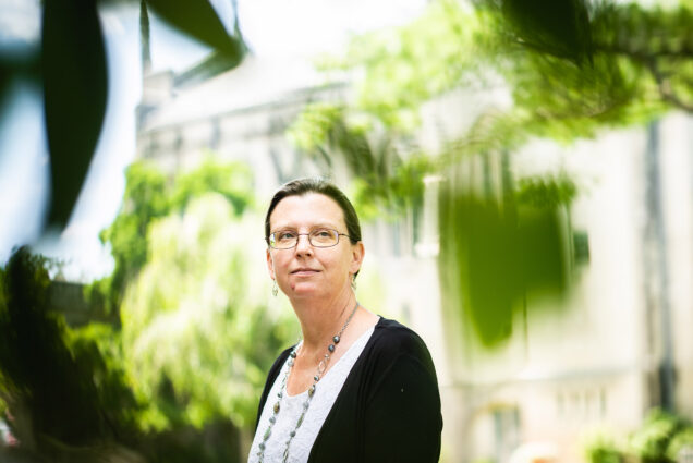 Photo: STH professor Rebecca Copeland poses for a photo on a bright green lawn. A white woman wearing glasses, a white blouse, black cardigan, and long beaded necklace, stands with hands placed in front of her. Foreground leaves frame her and an old church-like building is shown in the blurred background.