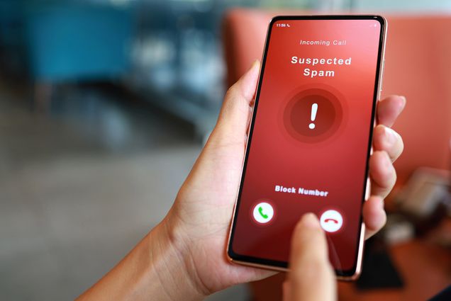 Photo: A hand shows the display of a cellphone screen. Screen shows text that reads "Suspected Scam" on a red background as the other hand's finger moves to click the decline button.