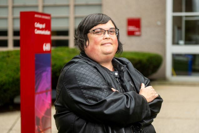 Photo: Joan Donovan, a white woman with short, silver hair and wearing a black top, faux leather jacket, and glasses, poses in front of the BU College of Communication building. Behind her, a red sign reads "College of Communication".