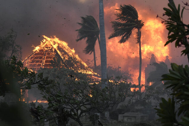 Photo: The hall of historic Waiola Church in Lahaina and nearby Lahaina Hongwanji Mission are engulfed in flames along Wainee Street. A large pyramid-shaped building and surrounding palm trees and land are shown engulfed in flames. Large flames are seen in the background as the sky looms red and black.
