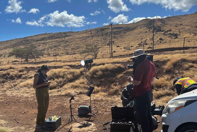 Photo: Melissa Adan of ABC News is shown reporting live from a roadside in Maui. A tan woman with black hair tied back and wearing a navy blue sleeveless blouse and khaki pants looks to her cellphone as a man wearing a large bucket hat, red shirt, and jeans holds a large TV camera in front of her. they stand on a dry, dirt road as fluffy clouds litter the blue sky.