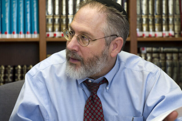 Photo: Dr. Michael A. Grodin, a white man with graying brown hair and beard, and wearing glasses, a light blue collared shirt, and brown tie looks to the left. He sits in front of a large bookcase filled with various reference books.