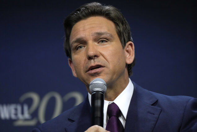 Photo: Ron DeSantis, a white man with short brown hair, speaks into a microphone in a close-up portrait. He is wearing a dark suit.