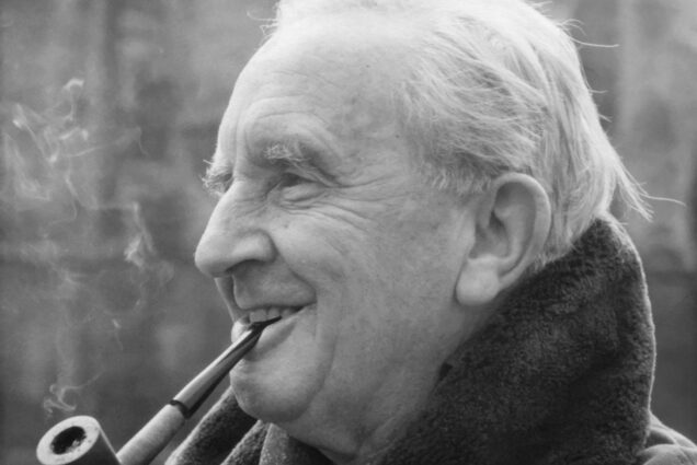 Photo: A black and white photo of J.R.R. Tolkien smoking a pipe. It hangs, leisurely, in his mouth and he smiles, amused, to something off camera. He wears a coat, fur-lined with an exposed and popped collar. His hair is short and white. The background is blurred, but he looks to be on a campus.